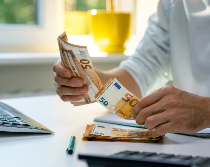 Osoba počítajúca 50-eurové bankovky pri stole, s klávesnicou, perom a hromádkou hotovosti a zvažuje, či použiť svoju kartu na nadchádzajúci nákup.