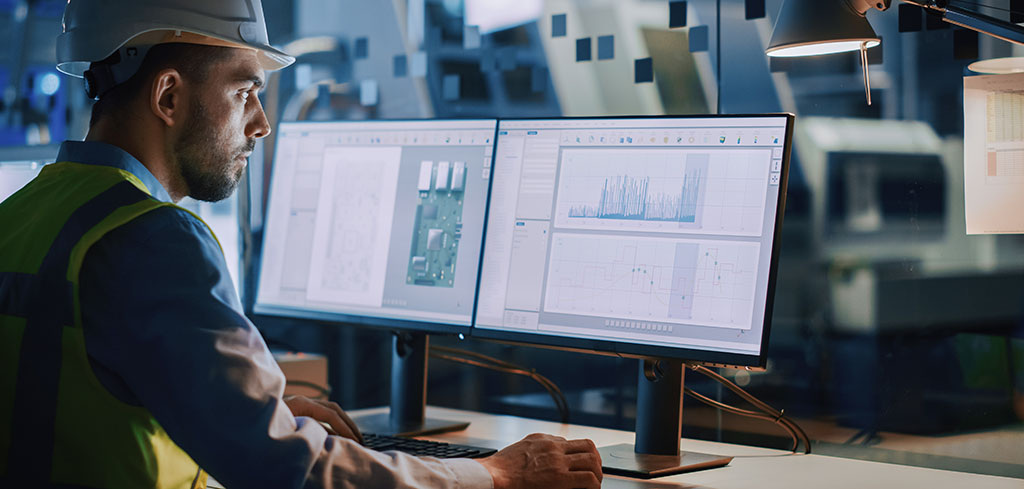 Muž v ochrannom odeve pracuje za stolom a analyzuje grafy údajov na dvoch počítačových monitoroch v priemyselnom prostredí so zameraním na dodržiavanie BOZP v prenajatých priestoroch.