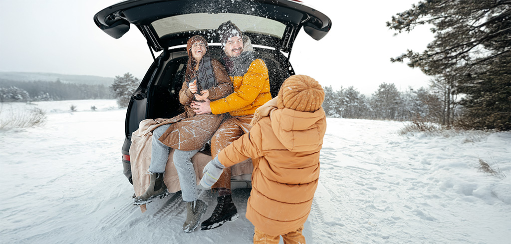 Dieťa hádže sneh na rodičov, ktorí sedia na zadnom sedadle auta v zasneženej krajine, zdieľajú smiech a chvíle, ktoré robia zimnú dovolenku nezabudnuteľnou - presne také spomienky si môžete vychutnať, keď viete ako uplatniť rekreačný poukaz.