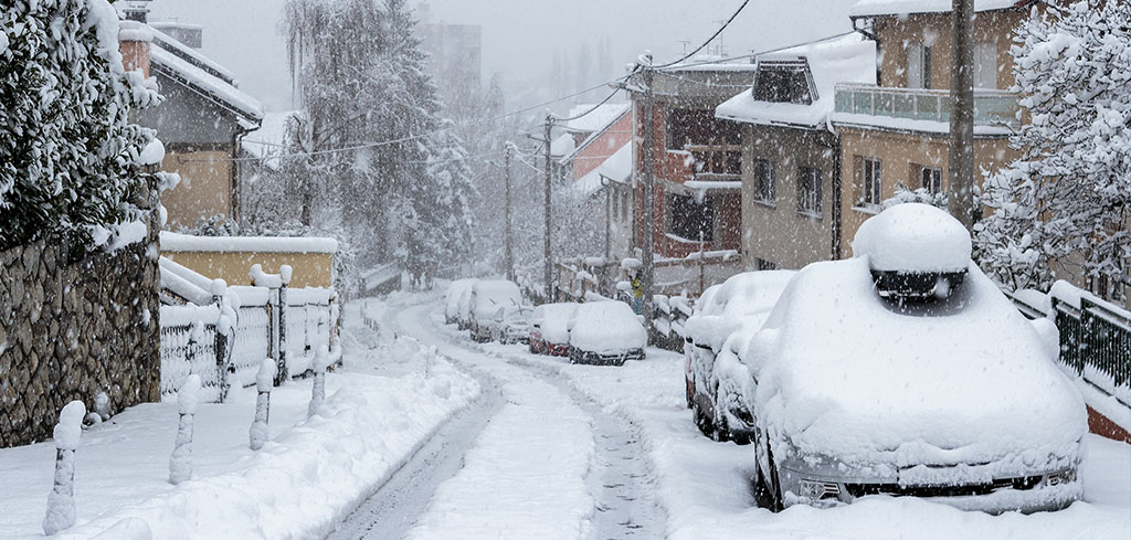 Ulica zasnežená ťažkým snehom, so zaparkovanými autami a domami zasypanými snehom - pripomína dôležitosť poistenia majetku a auta v zime.