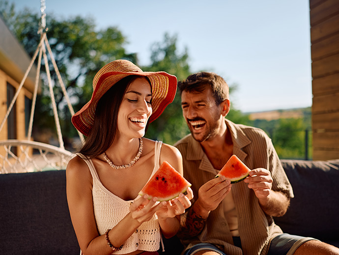 Smejúci sa pár sedí vonku počas slnečného dňa a pochutnáva si na plátkoch melóna.