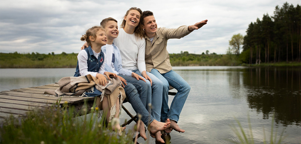 Rodina sedí na móle pri jazere, teší sa zo spoločne stráveného času, v pozadí sú stromy a oblačná obloha.