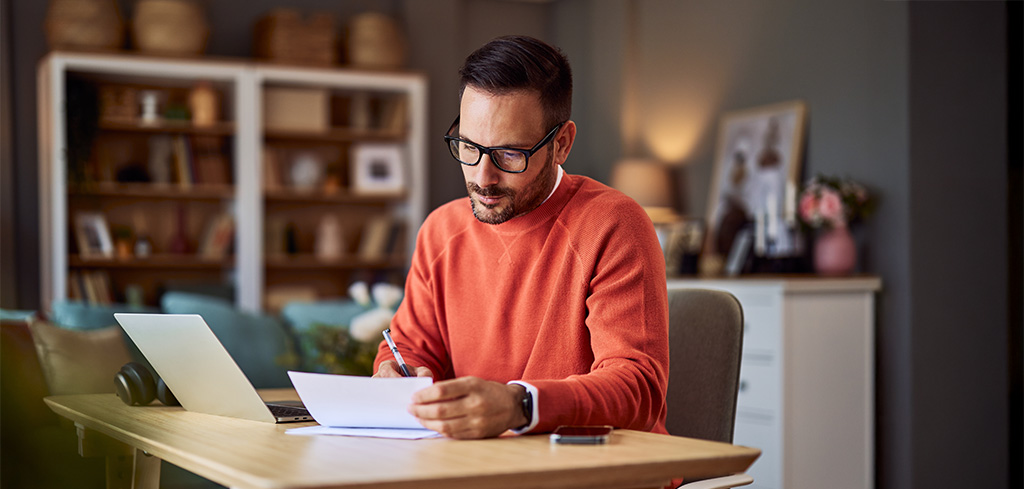Muž v oranžovom svetri pracuje za stolom s notebookom, drží papiere a píše poznámky počas kontroly spolupráce so živnostníkmi.