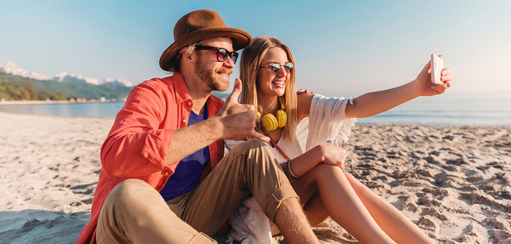 Šťastný pár si spoločne robí selfie na piesočnej pláži, obaja majú na sebe slnečné okuliare – užívajú si dovolenku s Booking za benefity.