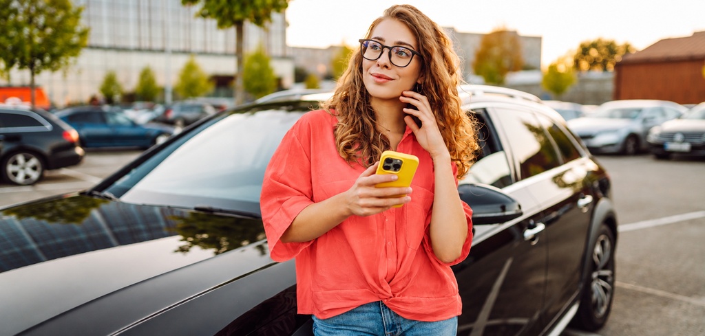 Mladá žena v okuliaroch drží v ruke smartfón, opiera sa o auto na parkovisku a vyberá si PZP.